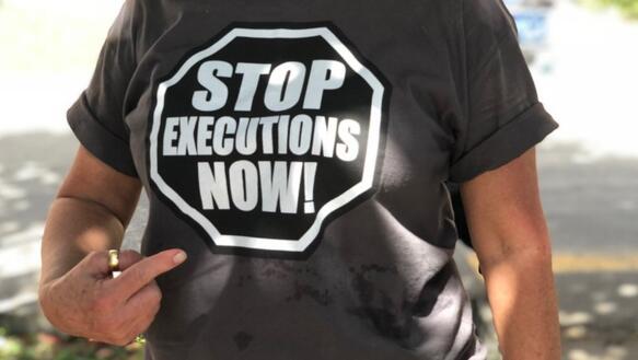 Aufnahme einer Person an einem sonnigen Tag, die mit ihrer rechten Hand auf ihr T-Shirt zeigt, auf dem steht: "Stob executions now!"