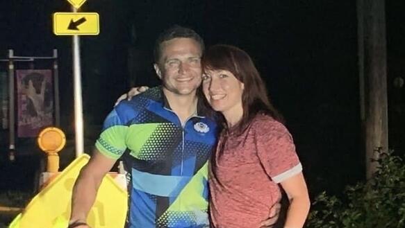 Das Foto zeigt Andriy Shepitsen, der in Sportkleidung hinter einem Fahrrad steht und in die Kamera lächelt. Neben ihm steht eine Frau, die ebenfalls lächelt. Sie umarmen sich gegenseitig. Das Foto wurde abends oder nachts aufgenommen vor einem Verkehrsschild.