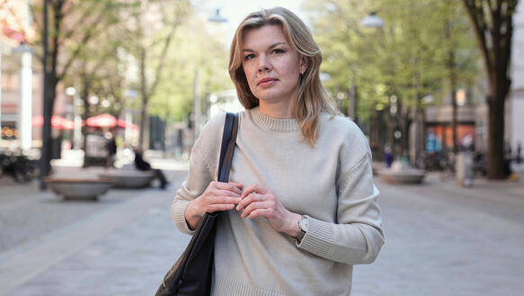 Eine junge Frau mit schulterlangem Haar trägt einen Strickpulli und eine Handtasche, sie steht draußen in einer Fußgängerzone.