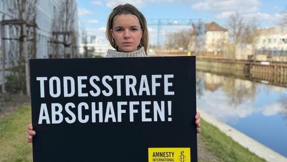 Das Foto zeigt eine junge Frau. Sie hält ein Schild mit Amnesty-Logo vor sich. Auf dem Schild steht: "Todesstrafe abschaffen". Links neben ihr steht ein Gebäude. Rechts von ihr ist ein Gewässer.