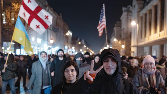 Demonstranten auf einer Straße in der Kälte, sie tragen Jacken, Schals, Mützen, schwingen Fahnen, ein Mann zeigt das Victory-Zeichen mit seinen Fingern.