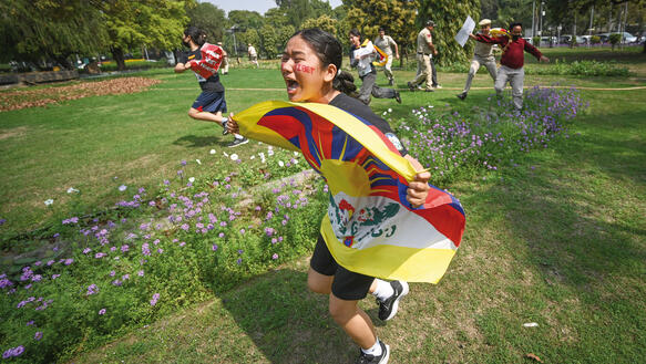 Eine Aktivistin rennt schreiend über eine Rasenfläche eines Parks, während sie eine tibetische Flagge vor ihrem Körper ausgebreitet hält; indische Polizeibeamte versuchen im Hintergrund andere Aktivisten festzuhalten.