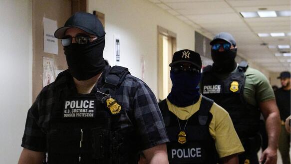 Das Foto zeigt drei Männer mit Basecap, Sonnernbrillen und Skimasken, die ihr halbes Gesicht bis unter die Augen bedecken. Sie gehen hintereinander in einem Flur. Sie tragen Schutzwesten über ihrer zivilen Kleidung und unter anderem eine Pistole im Holster.