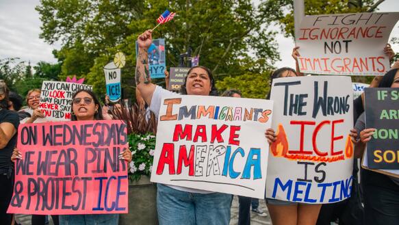 Das Foto zeigt mehrere Frauen bei einer Demonstration. Sie halten Schilder hoch. Auf einem steht: "The wrong ICE is melting". Auf einem anderen: "Immigrants make America".