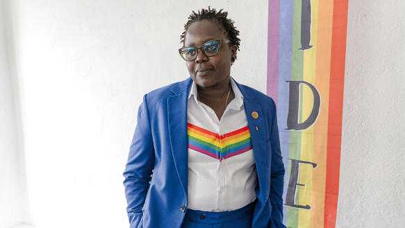 Eine Person im Anzug, darunter ein Hemd mit einem Keil in Regenbogenfarben, die Haare in kurzen Dreadlocks, eine Hornbrille aug, es ist die Pastorin Caroline Omolo