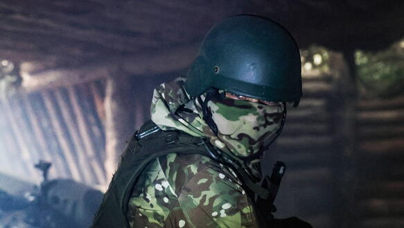 Das Foto zeigt einen maskierten Soldaten mit Helm und Schutzweste in einem Unterstand. Hinter ihm steht ein Geschützt.