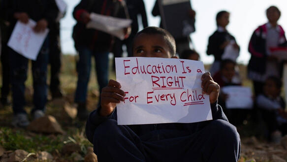 Eine Gruppe von Kindern steht mit Zetteln in der Hand an einem kleinen Hang. Ein Junge kniet vor ihnen und hält einen Zettel vor sich, auf dem steht: "Education is a right for every child".