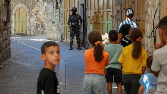 Eine Gruppe von Kindern in einer Gasse von hinten fotograftiert. Am Ende der Gasse steht ein maskierter Soldat mit Helm und Sturmgewehr. Nur eines der Kinder dreht sich um und blickt in die Kamera.
