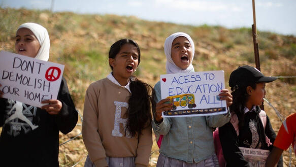 Vier Mädchen, von denen zwei Kopftücher tragen, steht vor einem kleinen Hang und rufen Slogans. Zwei von ihnen halten Schilder vor sich, auf denen unter anderem steht "Access to Education for all".
