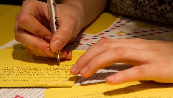 Nahaufnahme von Händen, die mit einem Kugelschreiber auf gelbem Papier einen Brief verfassen