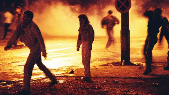 Menschen stehen auf einer von Feuer verrauchten nächtlich dunklen Straße, einige randalieren