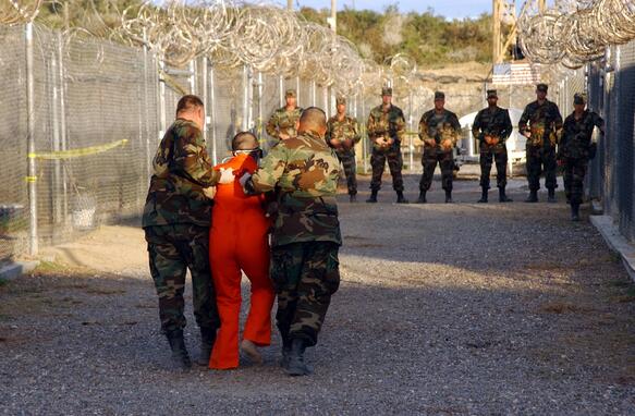 Das Bild zeigt Soldaten wie sie einen Mann in orangenem Overall abführen. Seitlich ist ein Zaun mit Stacheldraht zu sehen.
