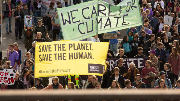 Eine protestierende Menschenmasse mit Bannern und Schildern, die friedlich demonstrieren. Auf dem gelben Amnesty-Banner steht "Save the planet, save the human".