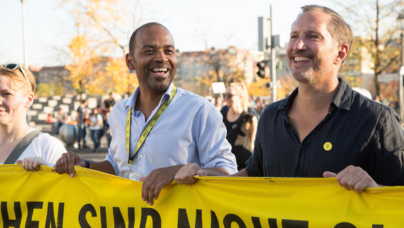 Zwei lächelnde Männer stehen hinter einem gelben Amnesty-Banner, den sie mit ihren Händen halten. Sie sind umgeben von anderen Menschen, die auch demonstrieren.
