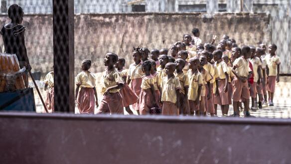 Durch ein Gitter fotografiert, zeigt das Bild eine Reihe von Schüler_innen in Schuluniform