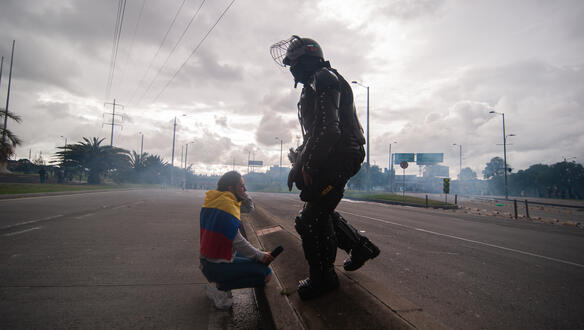 Eine Frau mit kolumbianischer Flagge auf einer Straße in der Hocke, vor ihr ein Polizist.