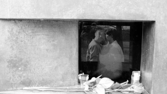 Das Bild zeigt eine Öffnung in einer Skulptur, davor liegen Blumen, man sieht ein Bild, auf dem sich zwei Menschen küssen