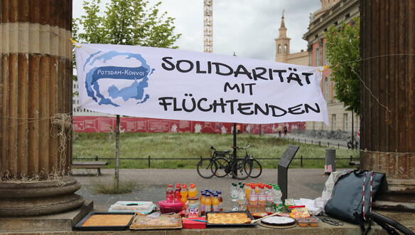 Das Foto zeigt ein Banner, das circa einen Meter über dem Boden zwischen zwei Säulen gespannt ist. Auf dem Banner steht: "Solidarität mit Flüchtenden".