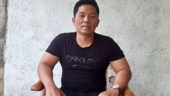 Heri Budiawan trägt ein T-Shirt und sitzt auf einem Stuhl mit beiden Armen auf den Lehnen und blickt ernst in die Kamera. 