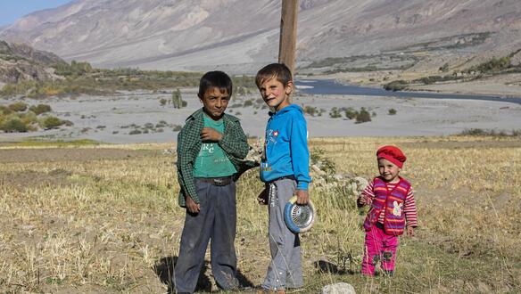 Drei Kinder unterschiedlichen Alters stehen in einer kargen Berg-Region, im Hintergrund ist neben wenigen kleinen grünen Pflanzen auch ein dünner Flusslauf zu erkennen. 
