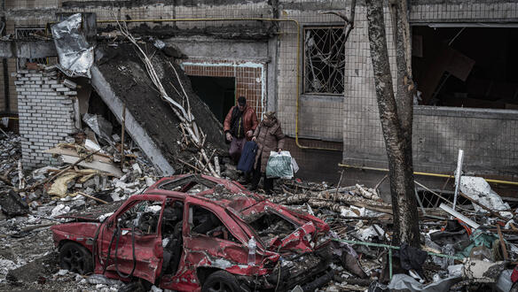 Das Bild zeigt zwei Menschen inmitten von Schutt, vor ihnen steht ein komplett zerstörtes Auto