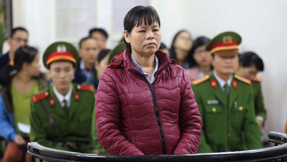 Das Foto zeigt  Cấn Thị Thêu stehend in einem Gerichtssaal. Sie hat die Hände gefaltet und Blick nach vorne. Hinter ihr sitzen Beamte in Uniform und Publikum.