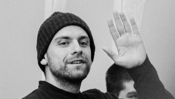 Das Foto zeigt Andro Chichinadze. Er trägt eine Strickmütze und lächelt und winkt mit seiner linken Hand.