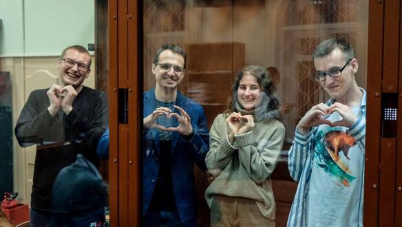 Das Foto zeigt die vier jungen Personen in einem Glaskasten. Ihre Hände sind mit Handschellen gefesselt. Sie formen ihre Hände zu einem Herzen und lachen in die Kamera. 