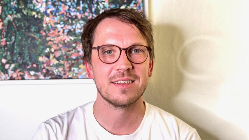 Porträt-Foto von Leonhard Kugler. Er trägt ein helles T-Shirt und eine Brille und lächelt in die Kamera.