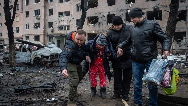 Drei Mäner stützten eine ältere Frau mit Gehstock vor einem zerstörten Gebäude.
