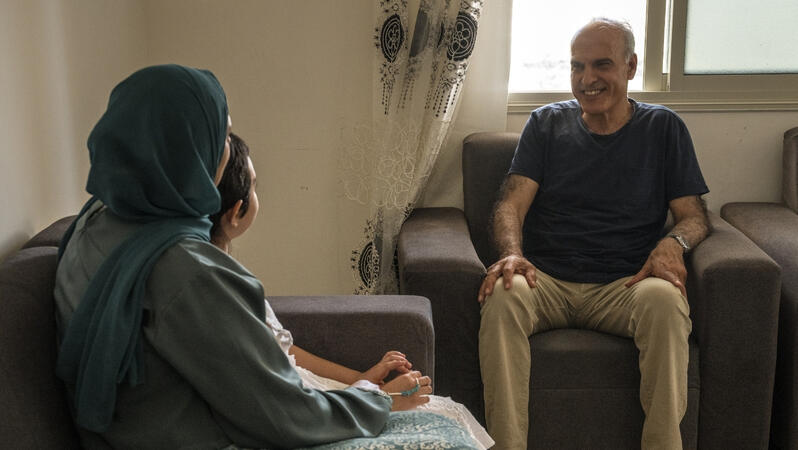 Khaled Quzmar sitzt lächelnd in einem Sessel, mit den Händen auf seinen Schenkeln. Zu seiner Rechten sitzen eine Frau mit Kopftuch und ein Mädchen auf einem Sofa. Er blickt sie lächelnd an.