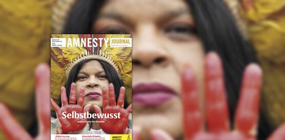 Eine indigene Frau trägt Lippenstift und einen Federkopfschmuck; sie streckt ihre Handflächen nach vorne aus, die mit roter Farbe eingefärbt sind. Das Foto trägt verschiedene Schriften, oben "Amnesty Journal", weiter unten "Selbstbewusst – Indigene fordern ihre Rechte ein"