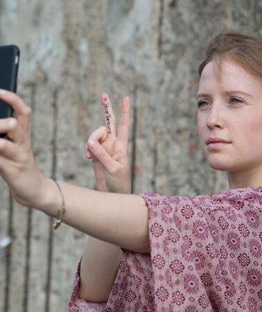 Eine junge Frau steht vor einer Mauer und macht das Victory-Zeichen. Dabei macht sie ein Selfie.