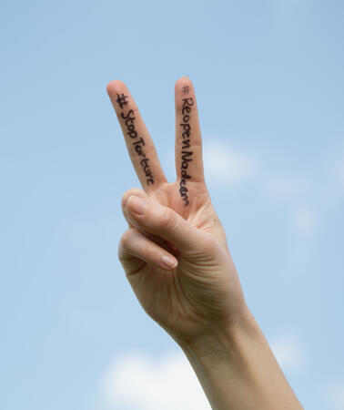 Eine Hand, die das Victory-Zeichen macht, vor blauem Himmel. Auf dem Zeigefinger und Mittelfinger steht #ReopenNadeem und #StopTorture