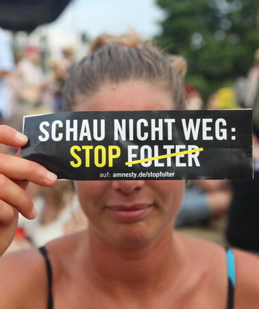 Eine junge Frau hält sich ein schmales Schild vor die Augen auf dem zu drauf steht: "Schau nicht weg. Stop Folter". Folter ist dabei durchgestrichen.