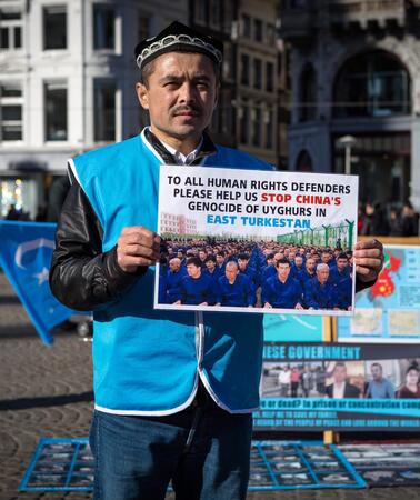 Auf einem Platz hält ein Mann in blauer Weste ein Schild, auf dem "To all human rights defenders please help us stop China's genocide of Uyghurs in East Turkestan" steht