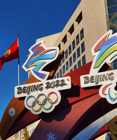 Das Bild zeigt ein Werbeschild der Olympischen Spiele vor einem Gebäude: Man sieht die Olympischen Ringe und den Schriftzug "Beijing". Dahinter sind chinesische Nationalflaggen zu sehen.