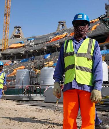 Zwei Menschen in Arbeitskleidung mit gelben Warnwesten auf einer Baustelle für ein Stadion. Ihre Gesichter sind verhüllt. 