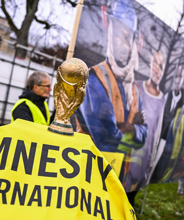Das Bild zeigt ein Amnesty-Banner, darauf ein Pokal und dahinter ein großes Plakat mit Menschen in Arbeitskleidung