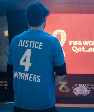 Das Bild zeigt zwei Personen von hinten, die auf einen Bildschirm schauen. Darauf steht "FIFA World Cup"