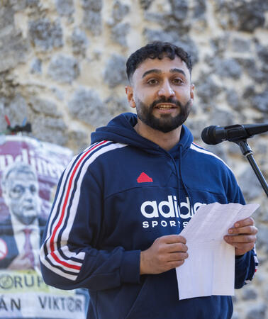 Géza Buzás-Hábel hält eine Rede. Vor ihm steht ein Mikrofon. Er trägt einen Kapuzenpullover. An einer Mauer hinter ihm hängt ein Protestplakat mit dem Foto von Ungarns Ministerpräsident Viktor Orbán.
