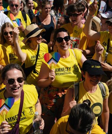 Das Foto zeigt Personen in Amnesty-Shirts inmitten einer großen Menschenmenge. Einige von ihnen halten herzförmige Papptafeln hoch mit Regenbogenfahne und dem Schriftzug Amnesty International.