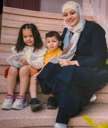 Das Foto zeigt Wesam Hamada mit einem Kopftuch auf einer Treppe sitzend. Neben ihr sitzen ihre Tochter und ihr Sohn. Sie hält ihren Arm um sie. Sie lächeln in die Kamera.