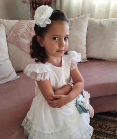 Hind Rajab steht in einem Zimmer vor einer Couch. Sie trägt ein Kleid und schaut mit verschränkten Armen in die Kamera.