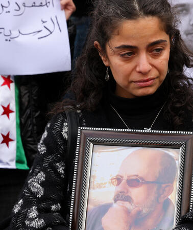 Eine weinende junge Frau hält ein gerahmtes Porträtfoto eines älteren Mannes vor der Brust. Im Hintergrund sind weitere Protestschilder mit Fotos und arabischer Schrift sowie eine Flagge der syrischen Opposition zu sehe
