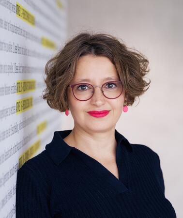 Porträtfoto von Wiebke Gülcibuk, die an eine Wand gelehnt in die Kamera lächelt. Sie trägt eine Brille.