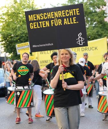Das Foto zeigt eine Frau an der Spitze eines Demonstrationszuges. Sie hält ein Schild hoch mit Amnesty-Logo und dem Slogan "Menschenrechte gelten für alle!". Hinter ihr spielt eine Trommelgruppe.