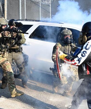 Bewaffnete Polizeikräfte gehen auf eine Person mit einer Gasmaske, einem Schild und einer Motorradjacke auf der "USA" steht zu.