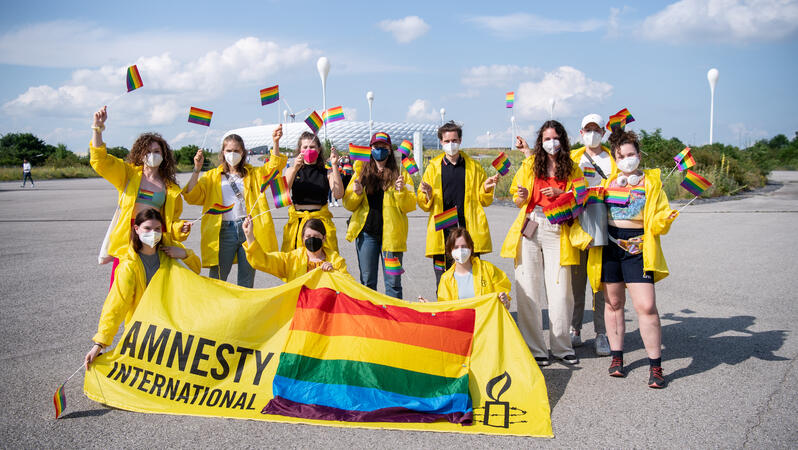 Menschen in gelben Jacken wedeln mit kleinen Regenbogenflaggen. Im Hintergrund ist die Allianz-Arena zu sehen, im Vordergrund halten mache der Menschen ein gelbes Banner mit einer großen Regenflagge drauf und mit der Aufschrift "Amnesty International". 