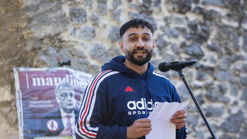 Géza Buzás-Hábel hält eine Rede. Vor ihm steht ein Mikrofon. Er trägt einen Kapuzenpullover. An einer Mauer hinter ihm hängt ein Protestplakat mit dem Foto von Ungarns Ministerpräsident Viktor Orbán.